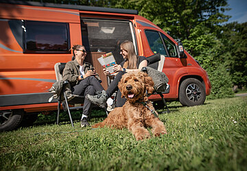 Zwei Personen und ein Hund sitzend vor einem organgefarbenen Camper 