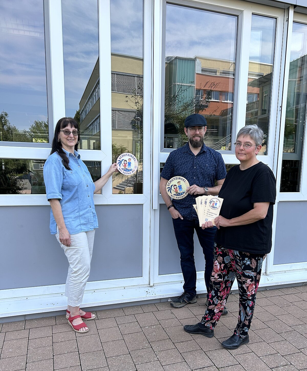 Drei Personen stehen vor einem Gebäude mit großen Glasfenstern und weißen Rahmen, auf dem Boden liegt ein gepflasterter Bereich; zwei Personen halten runde Schilder mit der Aufschrift „Familienstützpunkt“, eine Person hält mehrere Flyer in der Hand.