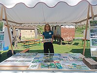 Person steht unter einem weißen Pavillon hinter einem Tisch mit zahlreichen Informationsbroschüren und Flyern, im Hintergrund sind Bänke, ein Baum und ein alter rostiger Wagen zu sehen.
