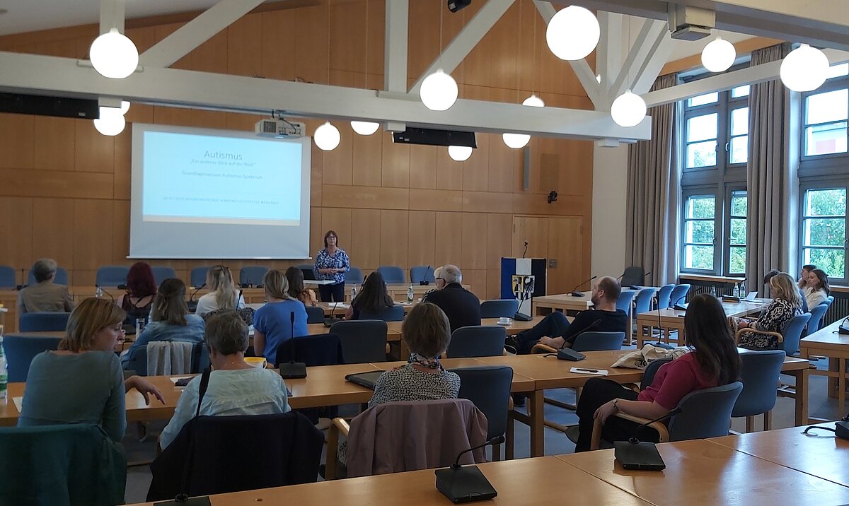 Konferenzraum mit Holzvertäfelung und großen Fenstern, mehrere Personen sitzen an Tischen und hören einer Vortragenden zu, die vor einer Leinwand steht, auf der eine Präsentationsfolie mit der Überschrift „Austausch“ und dem Text „Erfahrungen aus der Praxis – Herausforderungen und Lösungsansätze“ angezeigt wird.
