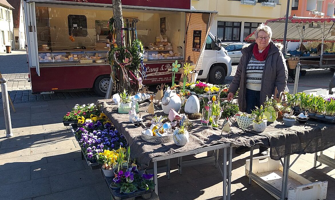Ein lebendiger Markttag in fränkischer Kulisse: Die Händlerin präsentiert liebevoll arrangierte Frühlingsblumen und Dekorationsartikel in weißen Keramiktöpfen – manche in Ei- oder Tierform – während im Hintergrund der rote Verkaufswagen „Lang & Laß“ mit Käse- und Backwaren lockt. Ein Bild voller Farben, Herzlichkeit und regionaler Vielfalt.