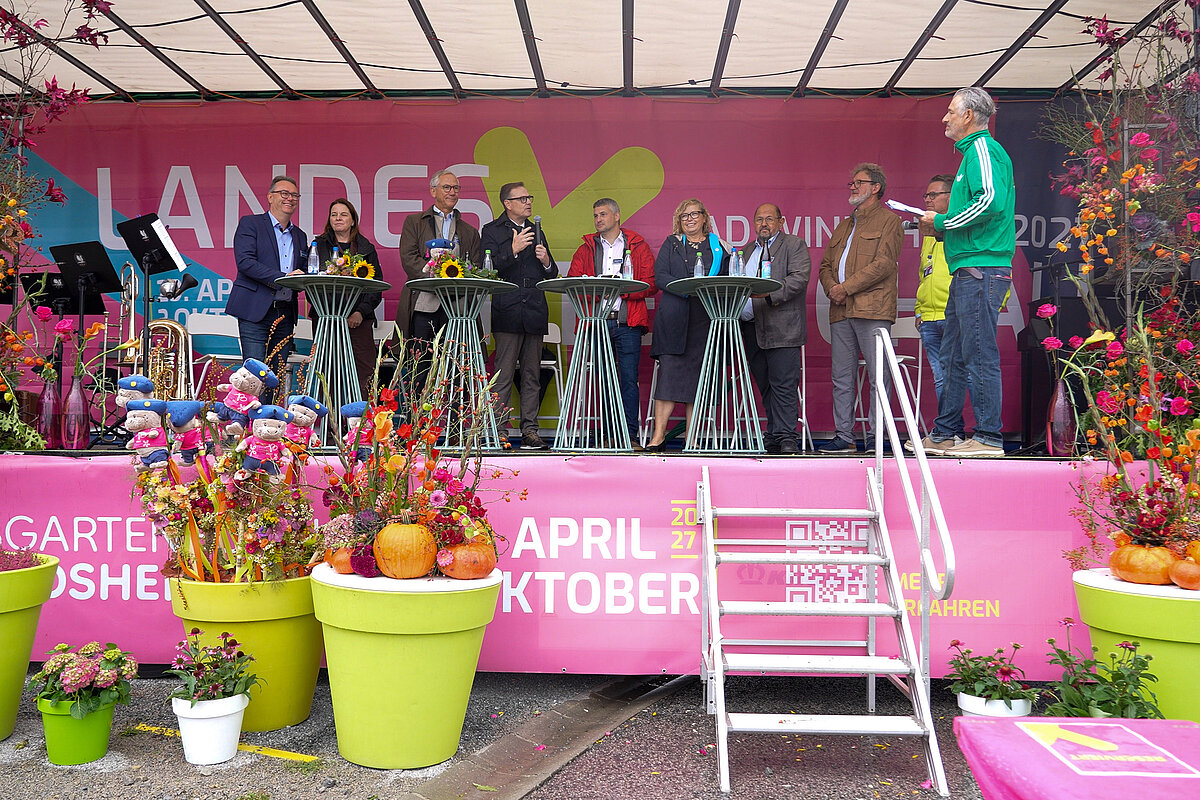 Prominente Persönlichkeiten auf einer pink gestalteten Bühne mit Plfanzen davor und einer Treppe
