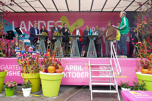 Prominente Persönlichkeiten auf einer pink gestalteten Bühne mit Plfanzen davor und einer Treppe 