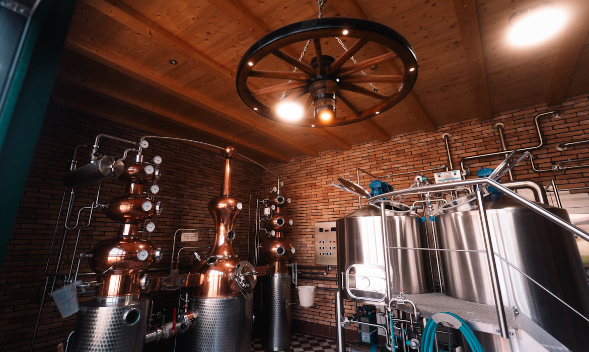 Innenraum einer Brennerei oder Brauerei mit gemauerten Wänden und Holzdecke – mehrere große Metalltanks und kupferne Brennblasen mit Ventilen und Rohrleitungen zeigen komplexe Verarbeitungstechnik; ein rustikaler Kronleuchter in Wagenradform hängt über dem schwarz-weiß gekachelten Boden und verbindet traditionelle Gestaltung mit moderner Produktionsausstattung.