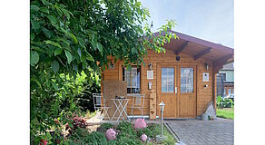 Holzhäuschen mit Sitzgruppe im grünen Garten – dekoriert mit rosa Schweinefiguren und Gartenleuchten, vermittelt ruhige, einladende Atmosphäre.