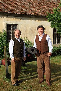 Zwei Personen stehen im Freien vor einem alten Gebäude mit Ziegeldach und verwitterter Fassade. Beide tragen weiße Hemden, braune Westen und braune Hosen. Neben ihnen befindet sich eine schwarze Kanone auf einem roten Sockel. Das Gebäude hat vergitterte Fenster und ist teilweise von grünem Bewuchs umgeben.