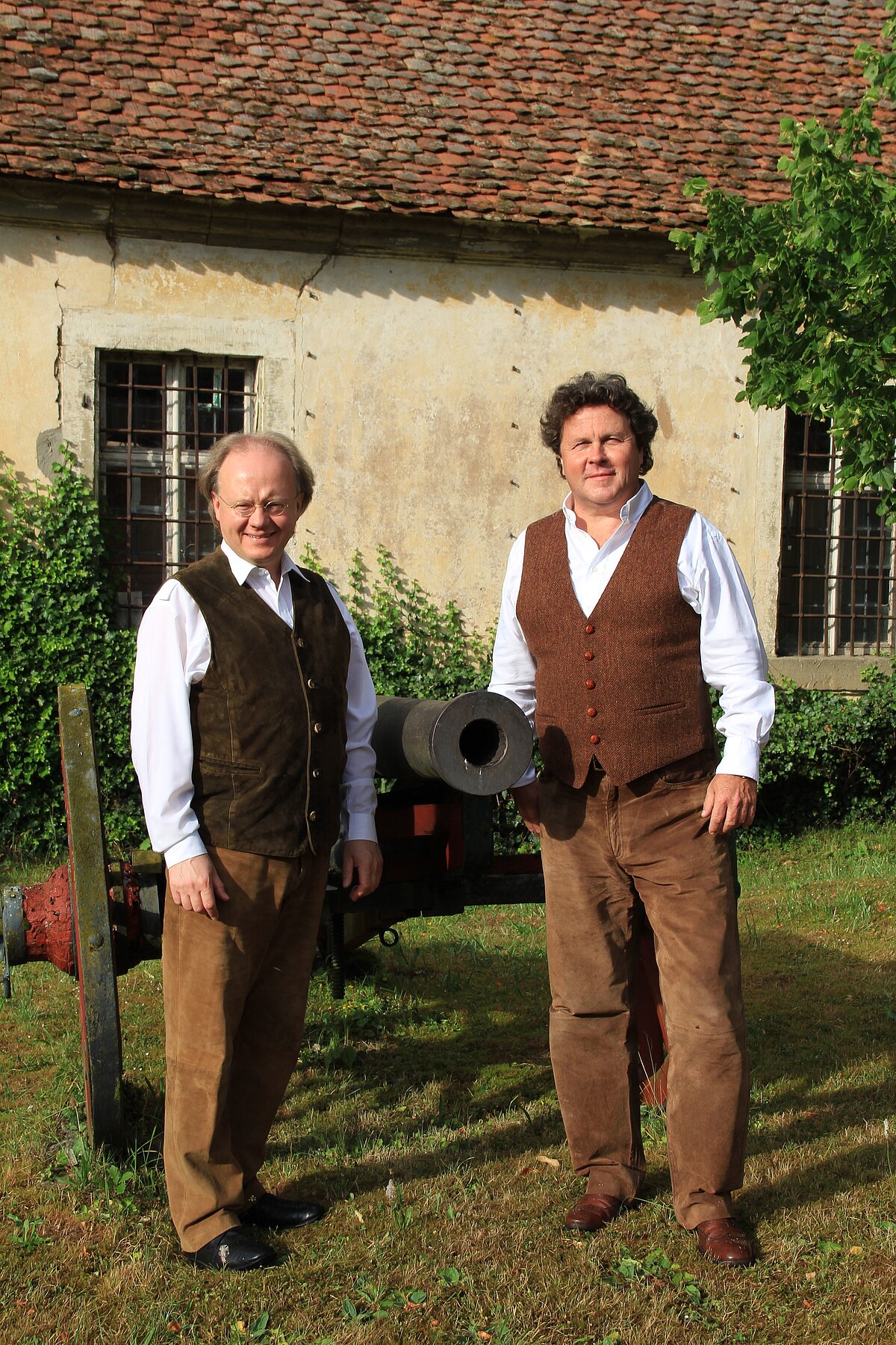 Zwei Personen stehen im Freien vor einem alten Gebäude mit Ziegeldach und verwitterter Fassade. Beide tragen weiße Hemden, braune Westen und braune Hosen. Neben ihnen befindet sich eine schwarze Kanone auf einem roten Sockel. Das Gebäude hat vergitterte Fenster und ist teilweise von grünem Bewuchs umgeben.