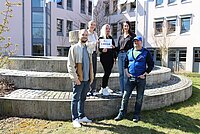 Fünf Personen stehen auf einer runden Steintreppe vor einem modernen Gebäude, eine Person hält ein Schild mit der Aufschrift „2.000 EUR“.