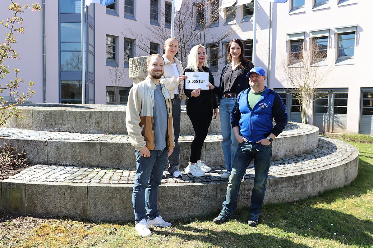 Fünf Personen stehen auf einer runden Steintreppe vor einem modernen Gebäude, eine Person hält ein Schild mit der Aufschrift „2.000 EUR“.