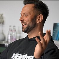 Mann mit Bart und schwarzem „KEMBRE“-Sweatshirt sitzt in einem modern eingerichteten Raum, lächelt und gestikuliert mit erhobenen Händen; im Hintergrund Vase mit Trockenblumen, Wasserflaschen und ein Stapel Magazine auf einem Schrank.