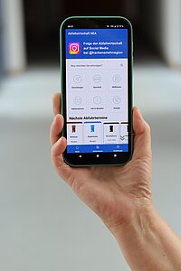 Eine Hand hält ein Smartphone, auf dem die App „Abfallwirtschaft NEA“ geöffnet ist. Die Benutzeroberfläche zeigt Symbole für Funktionen wie Erinnerungen, Standorte, Abfallarten, Rückruftermine, Infos & Aboservice und Kontakt. Darunter sind die nächsten Abfuhrtermine für Restmüll (Montag), Biomüll (Dienstag) und Papier (Mittwoch) aufgeführt.