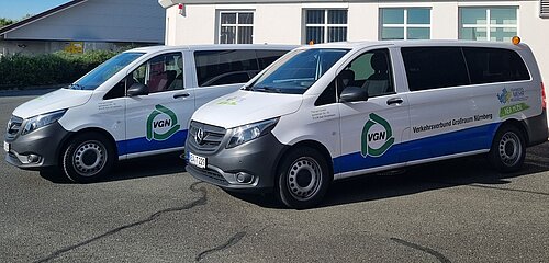 Zwei weiße Kleinbusse mit VGN-Logo und der Aufschrift „Verkehrsverbund Großraum Nürnberg“ stehen nebeneinander auf einem Parkplatz, beide mit blauer und grüner Beklebung sowie einem Hinweis auf „VGN Flexi“.