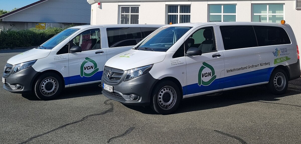 Zwei weiße Kleinbusse mit VGN-Logo und der Aufschrift „Verkehrsverbund Großraum Nürnberg“ stehen nebeneinander auf einem Parkplatz, beide mit blauer und grüner Beklebung sowie einem Hinweis auf „VGN Flexi“.