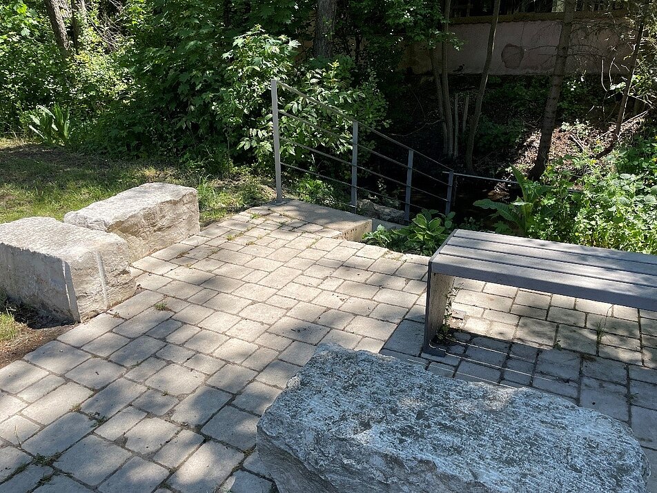 Kneippanlage im Leitenbach in Neuhof a.d. Zenn mit gepflastertem Aufenthaltsbereich, Sitzbank und Treppenzugang zum naturnahen Bachlauf.