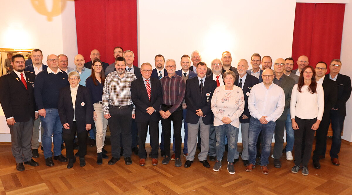 Gruppe von Menschen steht in einem Raum mit Holzfußboden und weißen Wänden, im Hintergrund zwei rote Vorhänge und eine Wandlampe, vorne eine Flasche Wasser auf einem Tisch sichtbar.