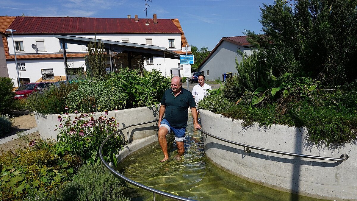 Zwei Personen gehen barfuß durch ein rundes Kneippbecken mit Handlauf, umgeben von blühenden Pflanzen und Wohnhäusern bei sonnigem Wetter.