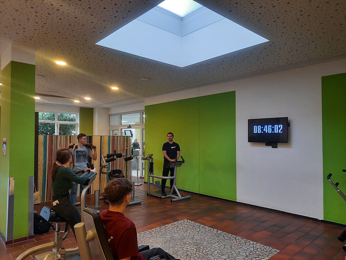 Ein Raum mit grün-weißen Wänden, Oberlicht und Fliesenboden. Mehrere Personen nutzen Trainingsgeräte, darunter ein Handkurbel-Ergometer. Eine Person filmt, eine steht am Laufband. 