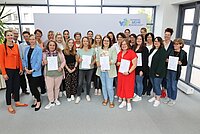 Große Gruppe von Personen steht in einem hellen Raum vor einem weißen Hintergrund mit dem Logo „Frankens MEHR Region“, mehrere Personen halten Zertifikate in den Händen, links und rechts sind große Fenster sichtbar.