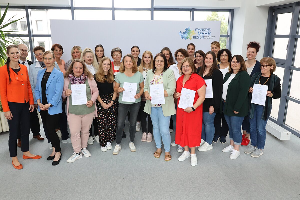Große Gruppe von Personen steht in einem hellen Raum vor einem weißen Hintergrund mit dem Logo „Frankens MEHR Region“, mehrere Personen halten Zertifikate in den Händen, links und rechts sind große Fenster sichtbar.