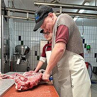 Zwei Personen in Schutzkleidung arbeiten in einer professionellen Fleischverarbeitungseinrichtung; eine Person schneidet ein großes Stück rohes Fleisch, während die andere daneben steht und zusieht.