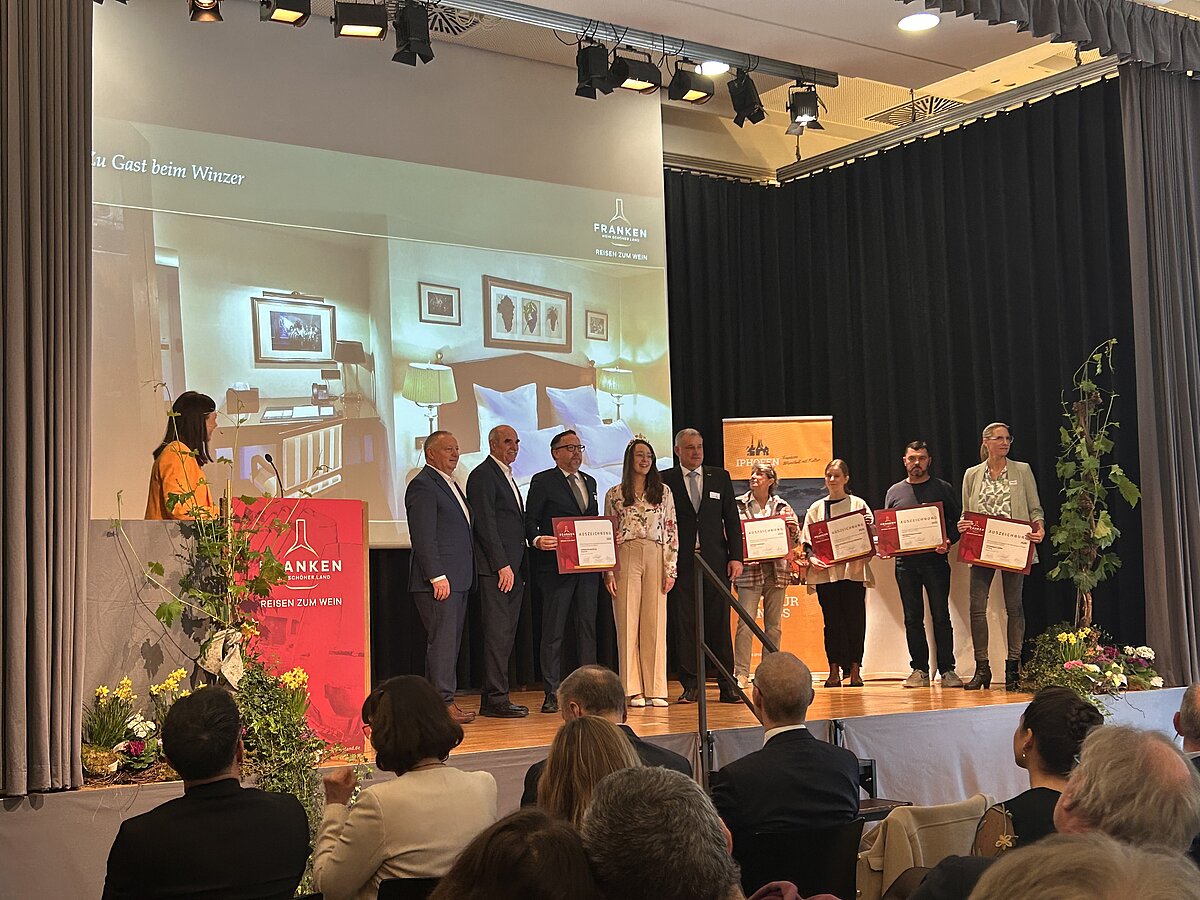 Gruppe von Personen auf Bühne bei feierlicher Zertifikatsverleihung – alle mit Urkunden in der Hand, im Hintergrund Projektion mit Schriftzug „PALMEN – Die Kunst der Wände“ und Innenraummotiven, Szene deutet auf Auszeichnung im Bereich Gestaltung oder Kunsthandwerk.