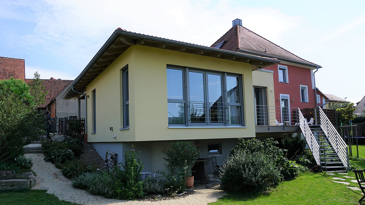 Zwei Wohnhäuser in gepflegter Umgebung: vorne ein modernes, gelb gestrichenes Gebäude mit Flachdach und Treppenaufgang, dahinter ein traditionelles rotes Haus mit Satteldach und weißen Fensterrahmen, umgeben von Garten und Sitzbereich.