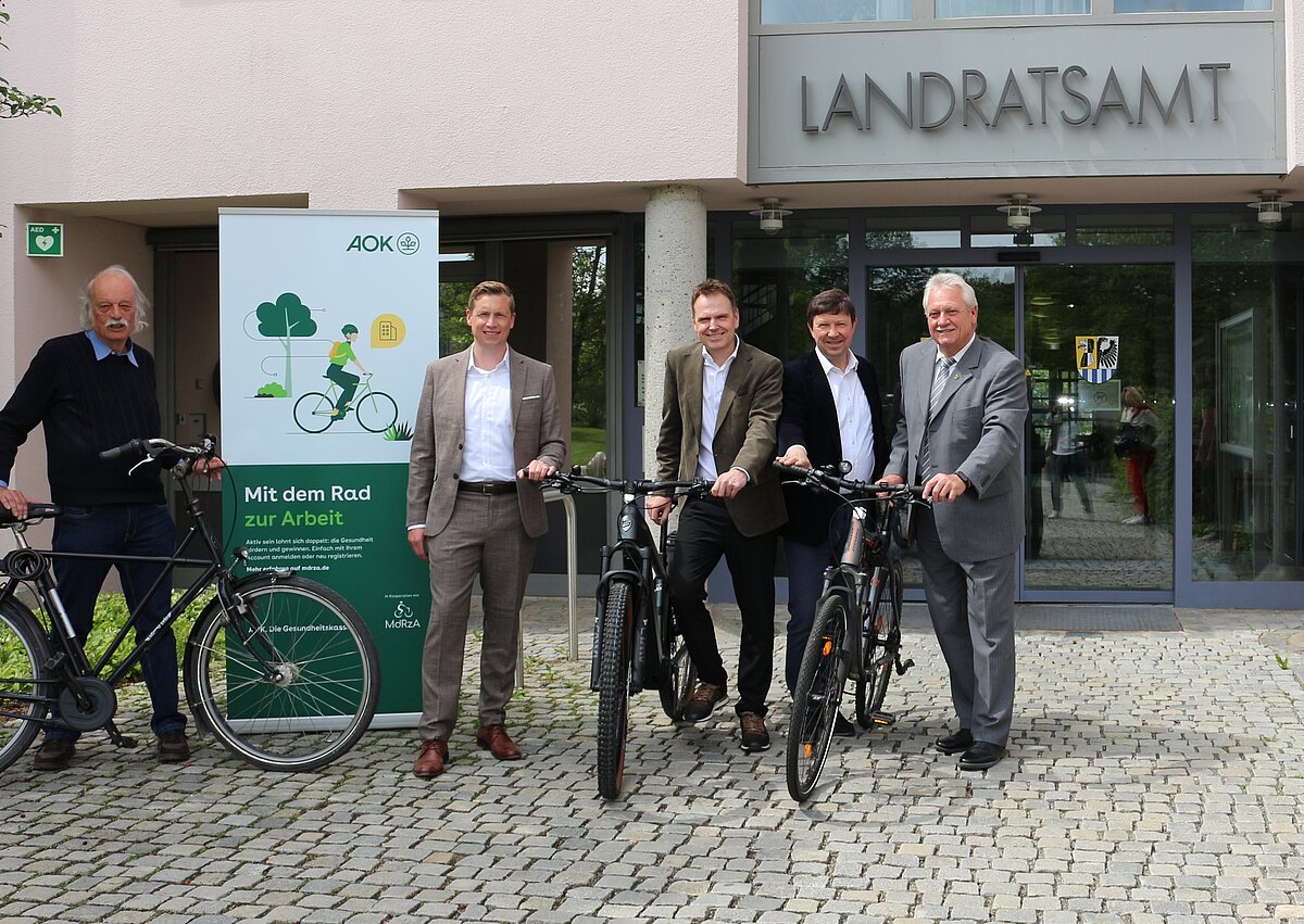 Fünf Personen stehen mit Fahrrädern vor dem Eingang des Landratsamts neben einem grünen Banner der AOK mit der Aufschrift „Mit dem Rad zur Arbeit“.