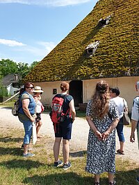 Mehrere Personen stehen vor einem historischen Gebäude mit einem sehr steilen, moosbedeckten Dach und kleinen Dachöffnungen, das eine zentrale Tür hat; im Vordergrund ein Kiesweg und Grasfläche, im Hintergrund Bäume unter blauem Himmel mit wenigen Wolken.
