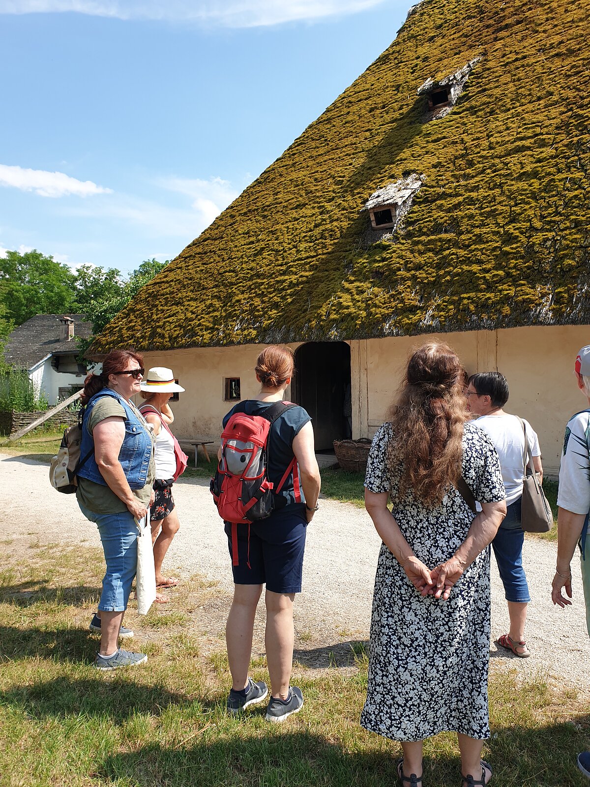 Mehrere Personen stehen vor einem historischen Gebäude mit einem sehr steilen, moosbedeckten Dach und kleinen Dachöffnungen, das eine zentrale Tür hat; im Vordergrund ein Kiesweg und Grasfläche, im Hintergrund Bäume unter blauem Himmel mit wenigen Wolken.