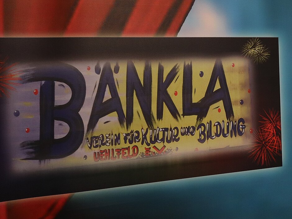 Ein Roter Vorgang der schrag durchs Bild geht und links unten von einer Hand gehalten wird, davor ein Schild mit der Aufschrift „Bänkla“