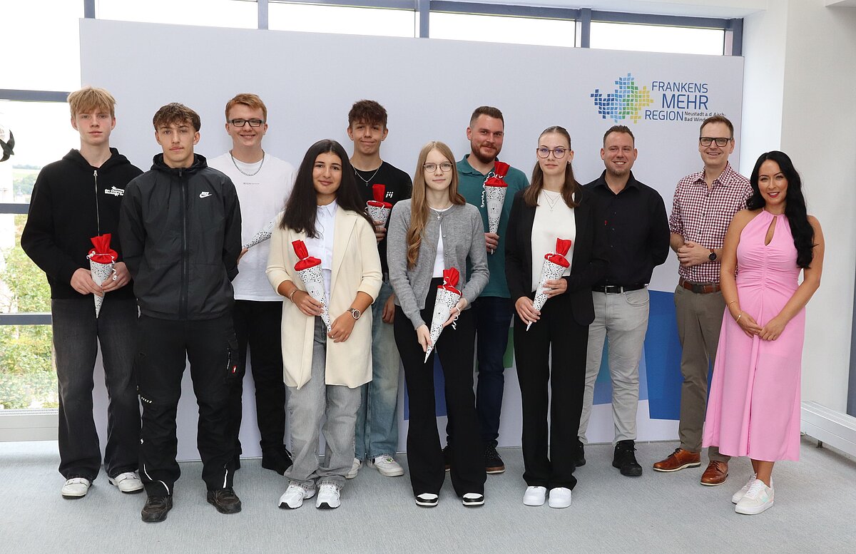 Elf Personen stehen vor einem weißen Hintergrund mit dem Logo „Frankens MEHR Region“ und halten jeweils eine rot-weiße Schultüte, rechts eine Person in einem pinken Kleid.
