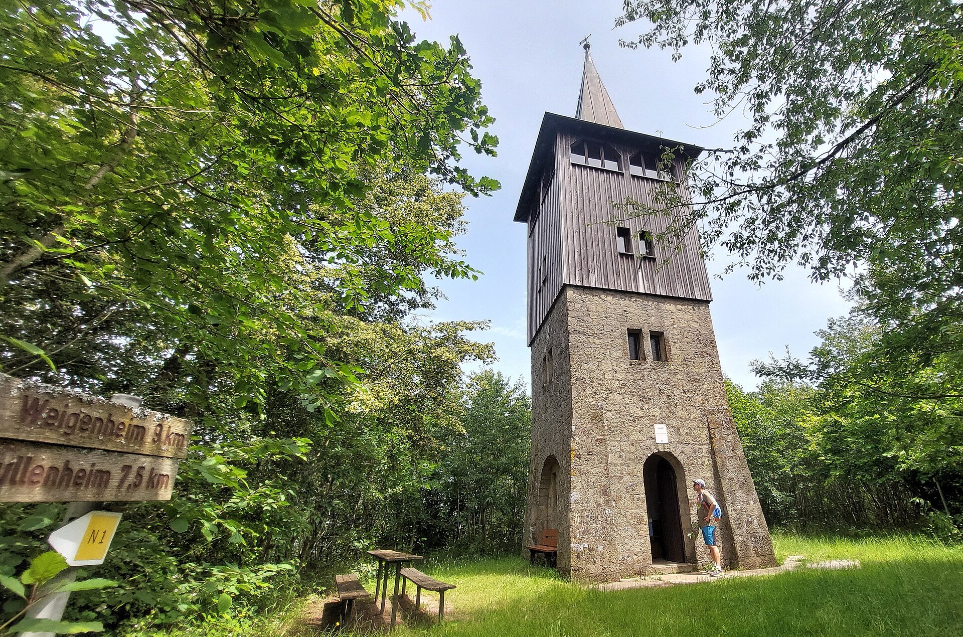 Links beginnt der Wald vom Turm, der Turm hat ein pyramidales Dach mit Holzlatten im obereen Bereich während die untere Hälfte aus Naturstein gemauert ist.