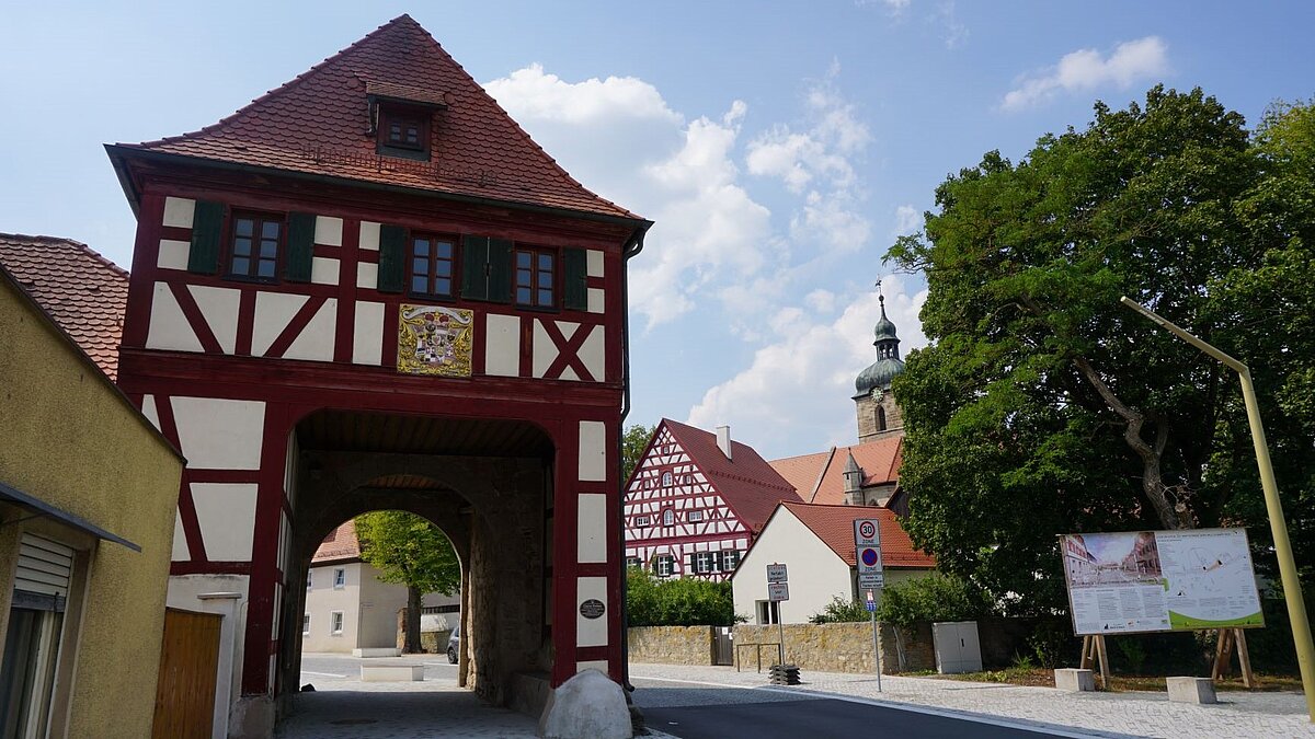 Historische Straßenansicht mit rot-weißem Fachwerk-Torhaus samt Wappen über dem Durchgang, flankiert von weiteren traditionellen Gebäuden und moderner Infotafel am Straßenrand.