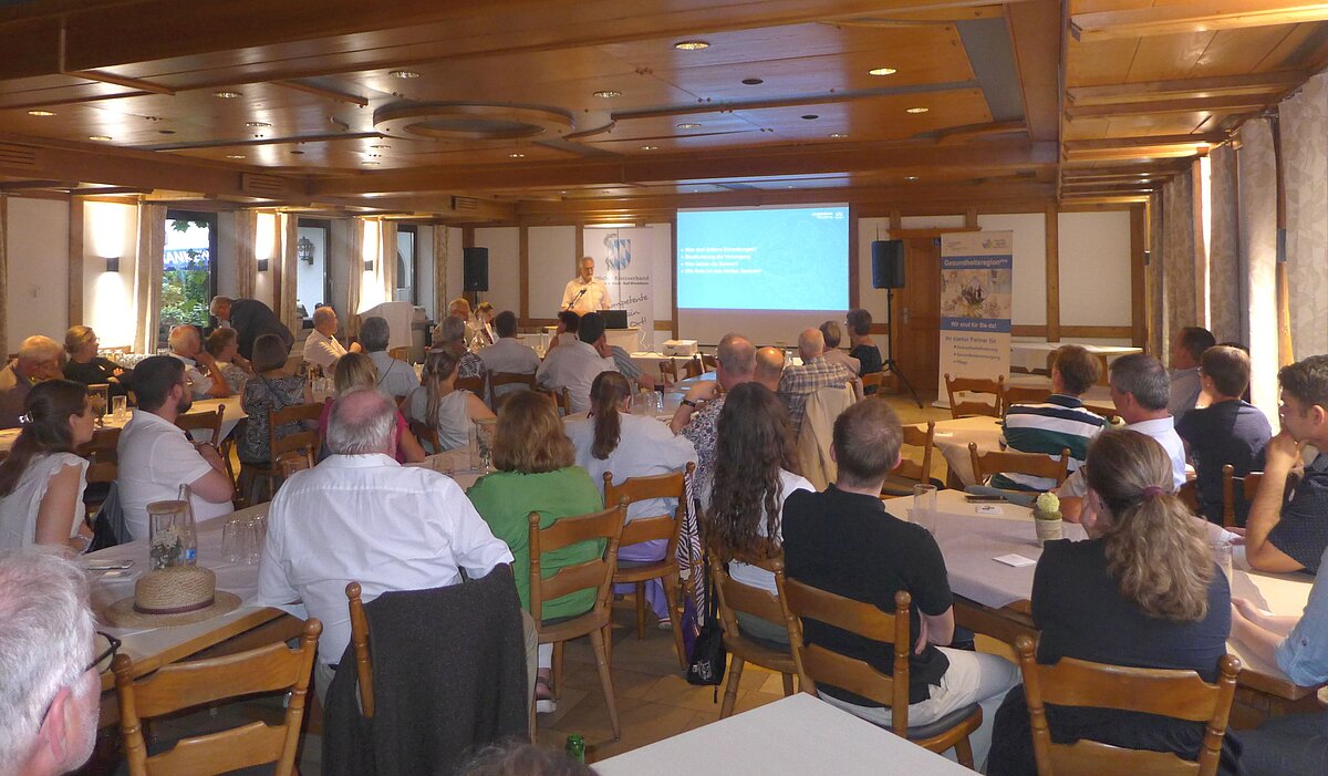 Mehrere Personen sitzen in einem holzvertäfelten Raum an runden Tischen und hören einer Präsentation zu, die auf einer Leinwand mit blauer Folie angezeigt wird; im Hintergrund stehen zwei Roll-Up-Banner, auf den Tischen liegen Gläser und Getränke.