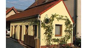 Kleines gelbes Haus mit rotem Ziegeldach und drei Holztüren – an der Fassade rankt eine Weinpflanze um Fenster und ein schildförmiges grünes Schild mit der Aufschrift „Weinbau Familie Mader“, davor Holzzaun und blühender Vorgarten, Szene vermittelt ländliche Idylle und familiären Weinbaubetrieb.