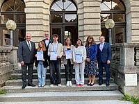 Acht Personen stehen vor einem historischen Gebäude mit Rundbogenfenstern. Vier in der Mitte halten Urkunden. Die Kleidung ist halbformell, das Setting wirkt wie eine Auszeichnungs- oder Abschlussveranstaltung.