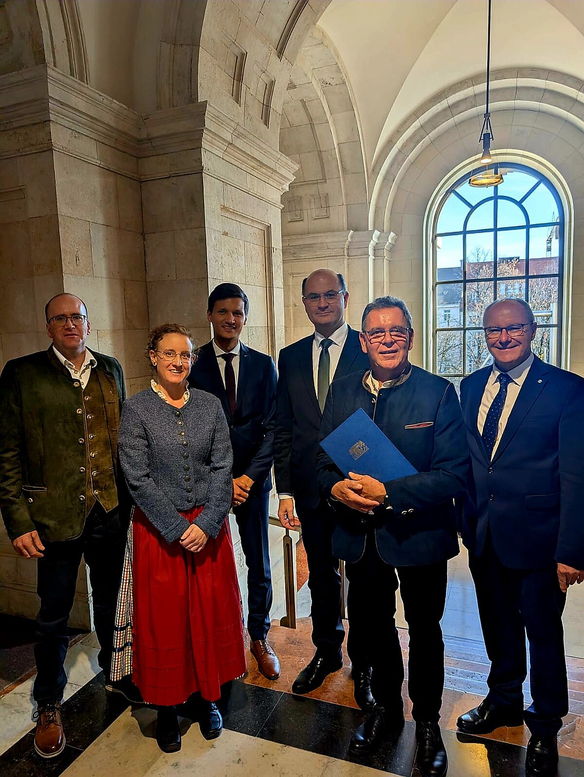 Sechs Personen stehen in einem Raum, eine davon hält eine Urkunde