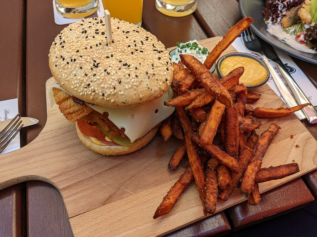 Karpfenburger mit Süßkartoffelpommes und zwei Dips, serviert auf einem Holzbrett in gemütlicher Außengastronomie. Die typische Panade und regionale Zubereitung machen ihn zu einer fränkischen Spezialität.