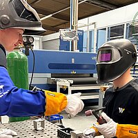 Zwei Personen in Schutzkleidung arbeiten gemeinsam an einem Schweißarbeitsplatz in einer Industrie- oder Ausbildungseinrichtung mit Maschinen und „JOB GUN“-Logo im Hintergrund.