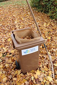 Braune Biotonne mit Laub und Rechen auf herbstlich bedecktem Boden – umweltbewusste Gartenpflege in Franken.