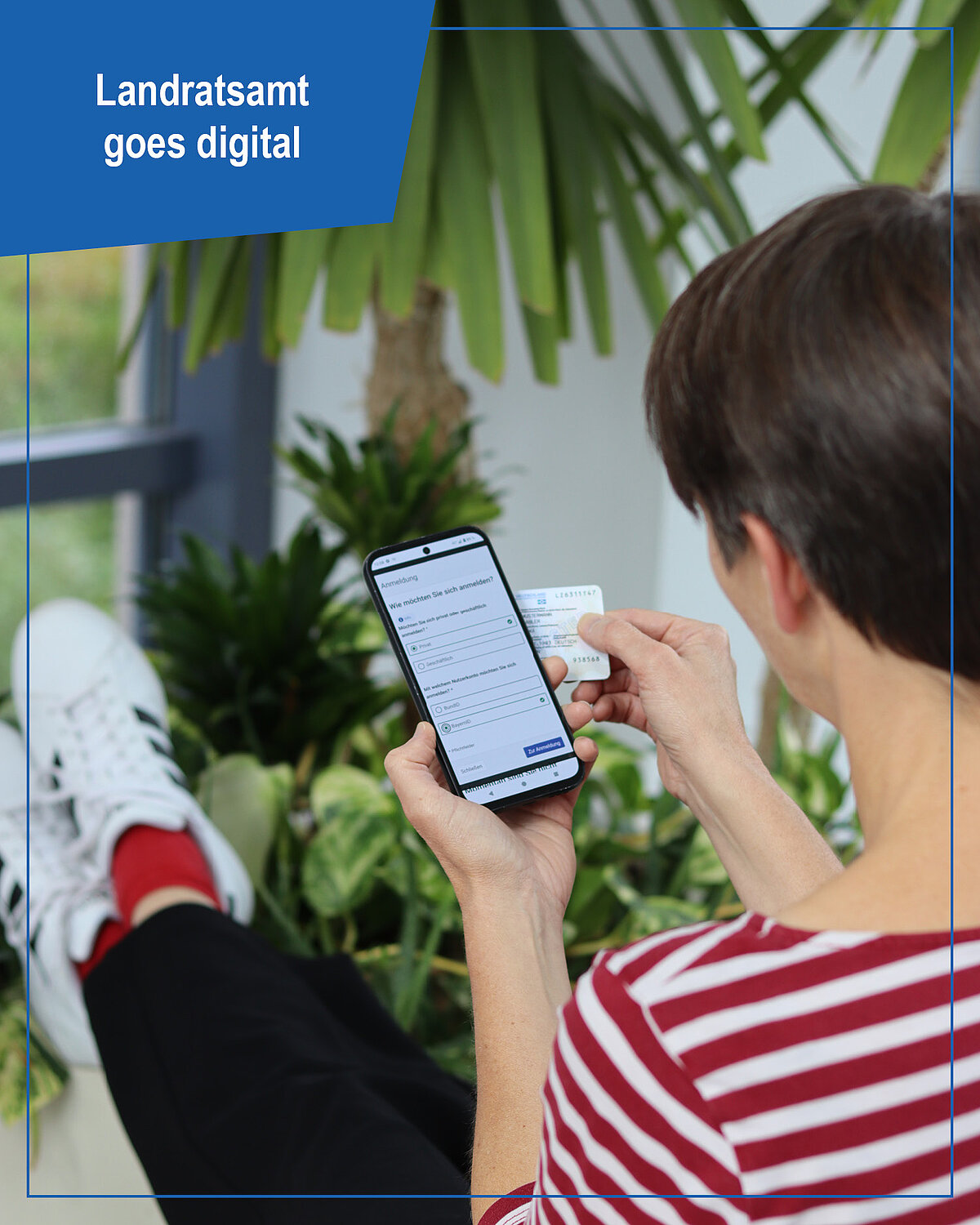 Eine Person in rot-weiß gestreiftem Shirt sitzt entspannt mit Smartphone und Karte in der Hand und nutzt ein digitales Formular; im Hintergrund sind grüne Zimmerpflanzen und ein Fenster zu sehen. Oben links steht „Landratsamt goes digital“.