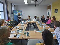 Gruppe von Personen sitzt um Tisch, eine Person steht am Flipchart