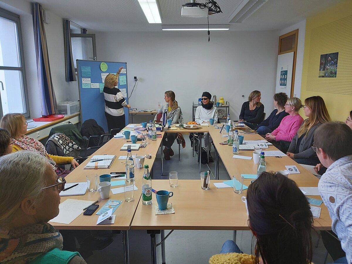Gruppe von Personen sitzt um Tisch, eine Person steht am Flipchart