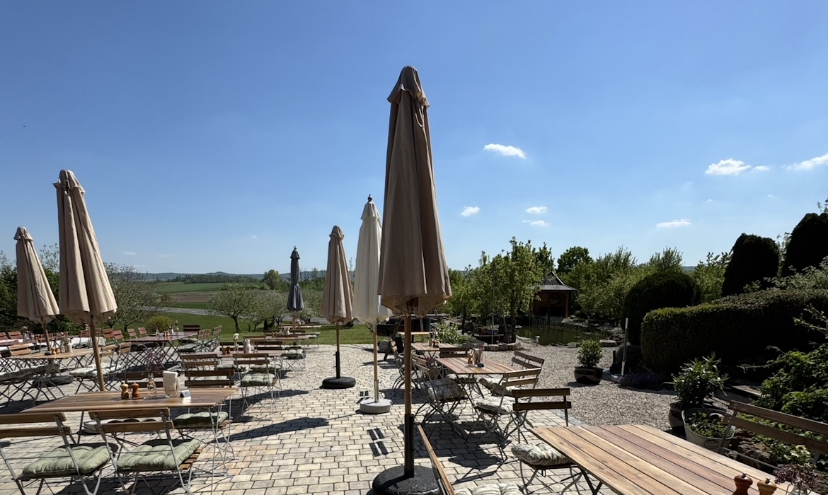 Gepflasterter Außenbereich eines Restaurants oder Cafés mit zahlreichen Holztischen und Metallstühlen – große beige Sonnenschirme spenden Schatten, im Hintergrund weite grüne Felder, Bäume und blauer Himmel mit wenigen Wolken; die Szene vermittelt eine ruhige, einladende Atmosphäre für entspanntes Essen im ländlichen Umfeld.