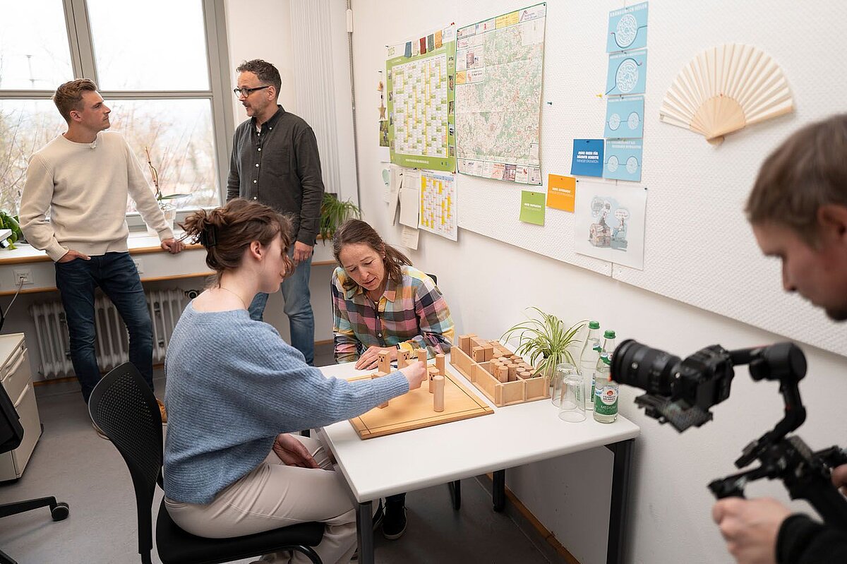 Fünf Personen befinden sich in einem Büro mit einem Tisch, auf dem ein Holzspiel oder Übungsgerät steht, zwei Personen sitzen und arbeiten daran, während zwei weitere danebenstehen und eine Person eine Kamera auf das Geschehen richtet; an der Wand hängen Karten, Notizzettel und ein Fächer.