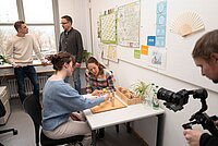 Fünf Personen befinden sich in einem Büro mit einem Tisch, auf dem ein Holzspiel oder Übungsgerät steht, zwei Personen sitzen und arbeiten daran, während zwei weitere danebenstehen und eine Person eine Kamera auf das Geschehen richtet; an der Wand hängen Karten, Notizzettel und ein Fächer.