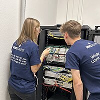 Zwei Personen in dunkelblauer Kleidung stehen vor einem offenen Serverschrank und arbeiten gemeinsam an Netzwerktechnik; auf ihren Rücken ist „AWS GRUPPE“ und IT-bezogene Schrift zu sehen.