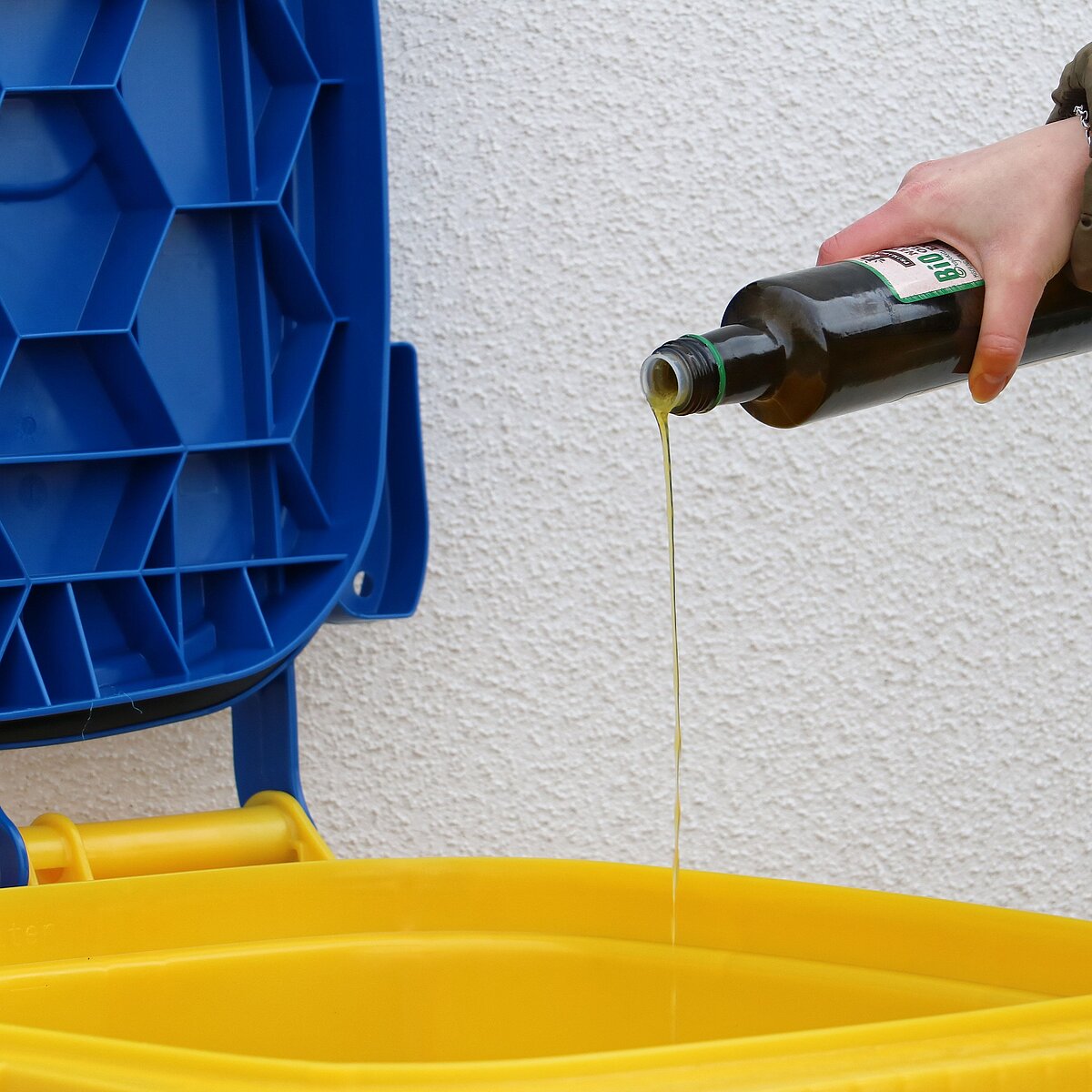 Eine Hand kippt eine grüne Glasflasche mit Öl über eine gelbe Tonne mit geöffnetem blauem Deckel, während das Öl in die Tonne fließt; im Hintergrund eine weiße Wand.