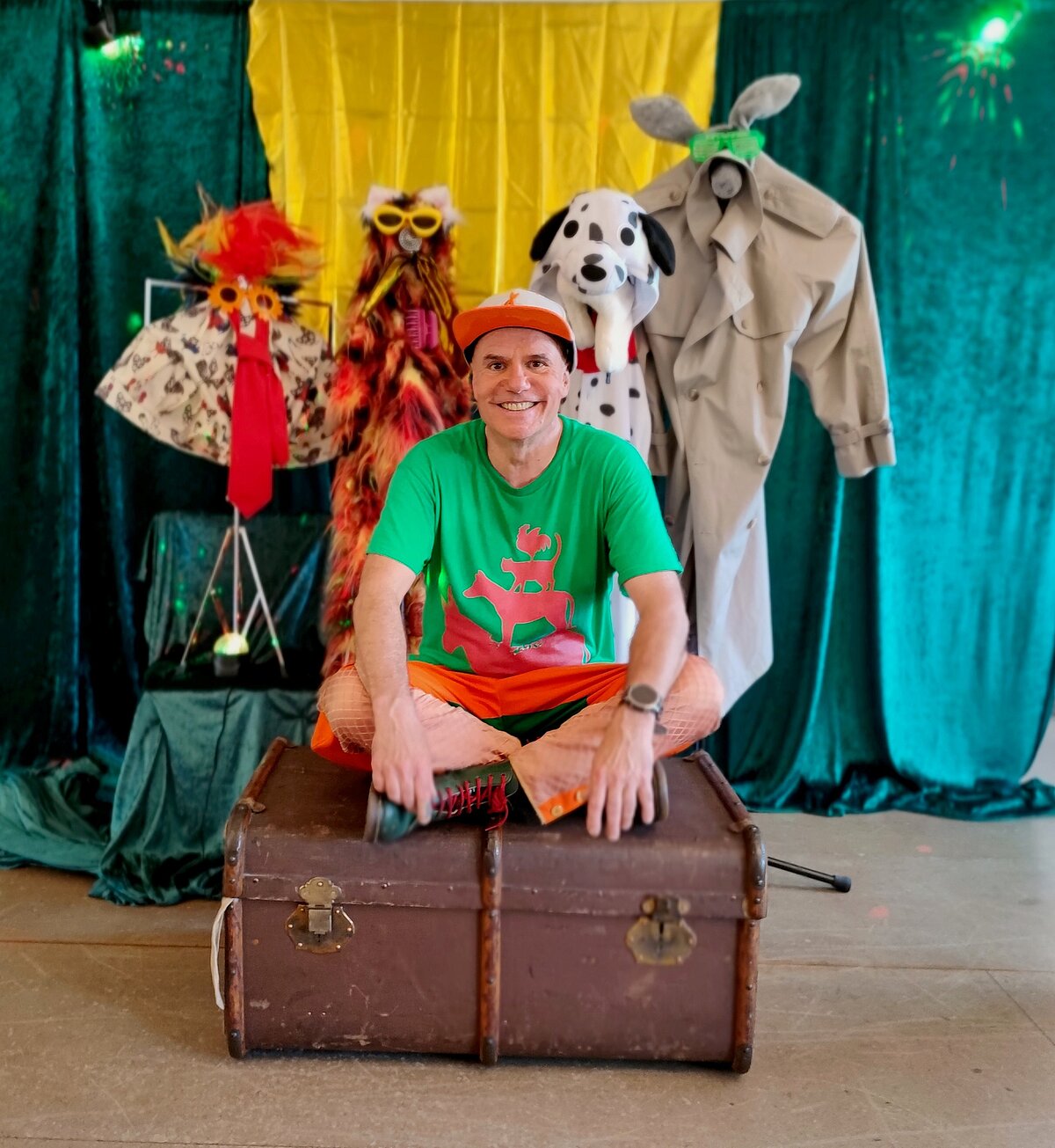 Person in grünem T-Shirt mit rotem Motiv und orangefarbener Hose sitzt auf einer großen braunen Truhe, im Hintergrund bunte Kulisse mit gelbem Stoff und drei dekorierten Figuren: eine mit rotem Federkleid und gelber Brille, eine Dalmatinerfigur und eine Figur mit grauem Mantel und Hasenohren.