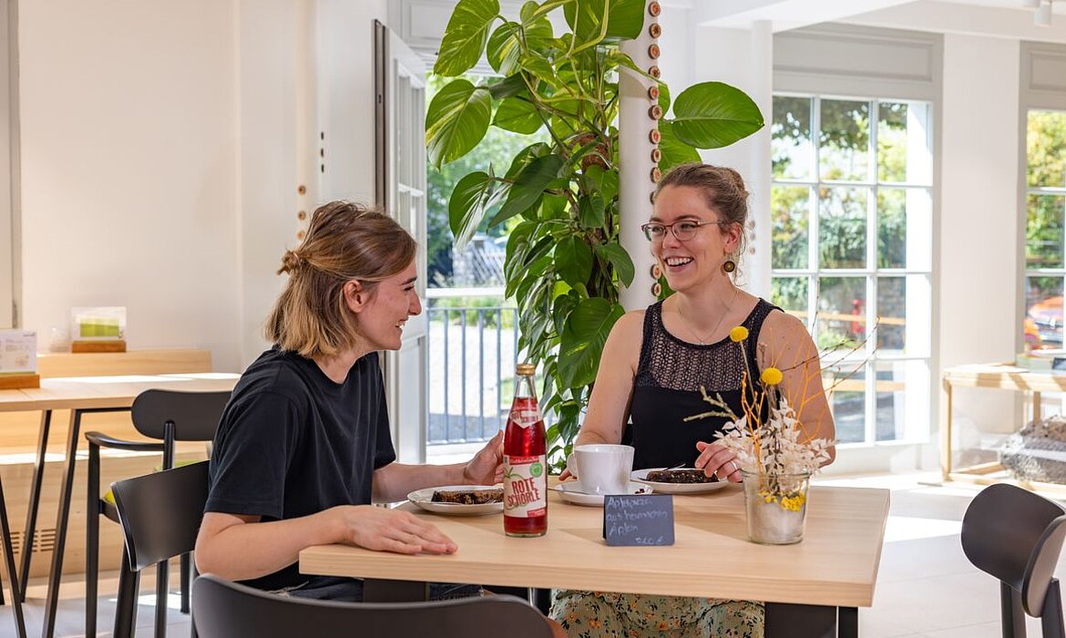 Zwei Frauen sitzen an einem hellen Holztisch in modernem, lichtdurchflutetem Innenraum – auf dem Tisch stehen Teller mit Speisen, eine rote „BIONADE“-Flasche, eine Tasse und ein kleines Schild mit „Afternoon Delight 4.50 €“; dekorative Vase mit gelben Trommelblumen und Trockenpflanzen, im Hintergrund große Grünpflanze, Fensterfront und Barhocker – Szene vermittelt entspannte, stilvolle Café-Atmosphäre.
